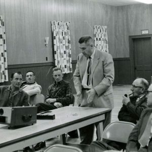 02-00-1961 Man at slide projector speaking with group of men#2-Website
