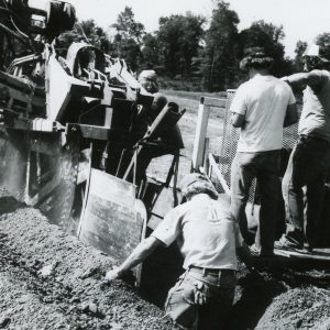 Man standing in ditch in front of digging machine. Three other men off to side. Photo by Robert Mills