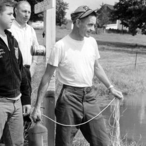 Man with jug beside pond as a man & boy look on .Photo by Robert Mills