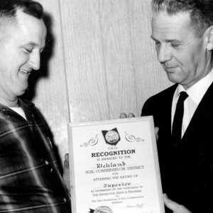 Two men holding Recognition plaque betwwen them & looking at it. Photo by Robert Mills
