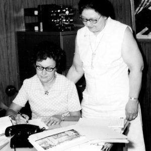 Woman at desk writing while a women standing looks on.Photo by Robert Mills