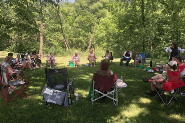 A large group of people sitting in soccer chairs in a circle in a clearing in a woods 