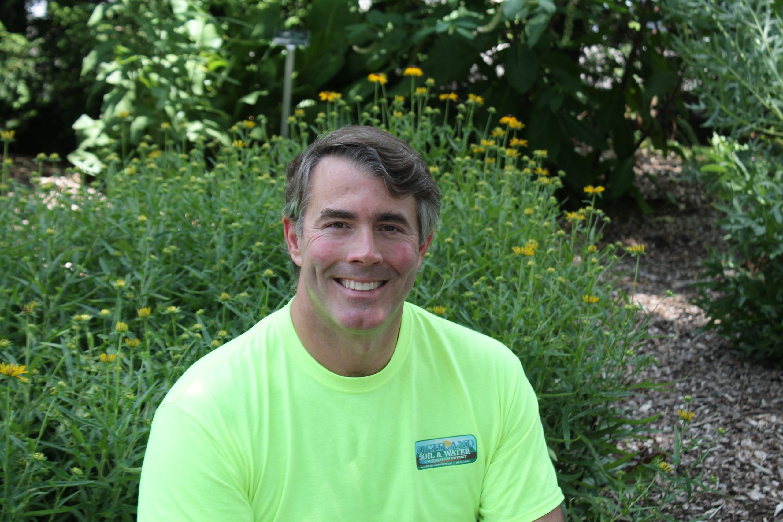 Matt seated in front of some yellow flowers that are just starting to bloom. He's wearing a Richland SWCD shirt and smiling for the camera.