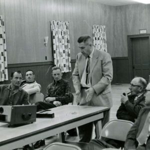 02_00_1961_Man_at_slide_projector_speaking_with_group_of_men_2_Website-5813