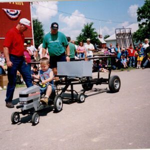 Little boy on small tractor as 2 men observe. Spectators in background